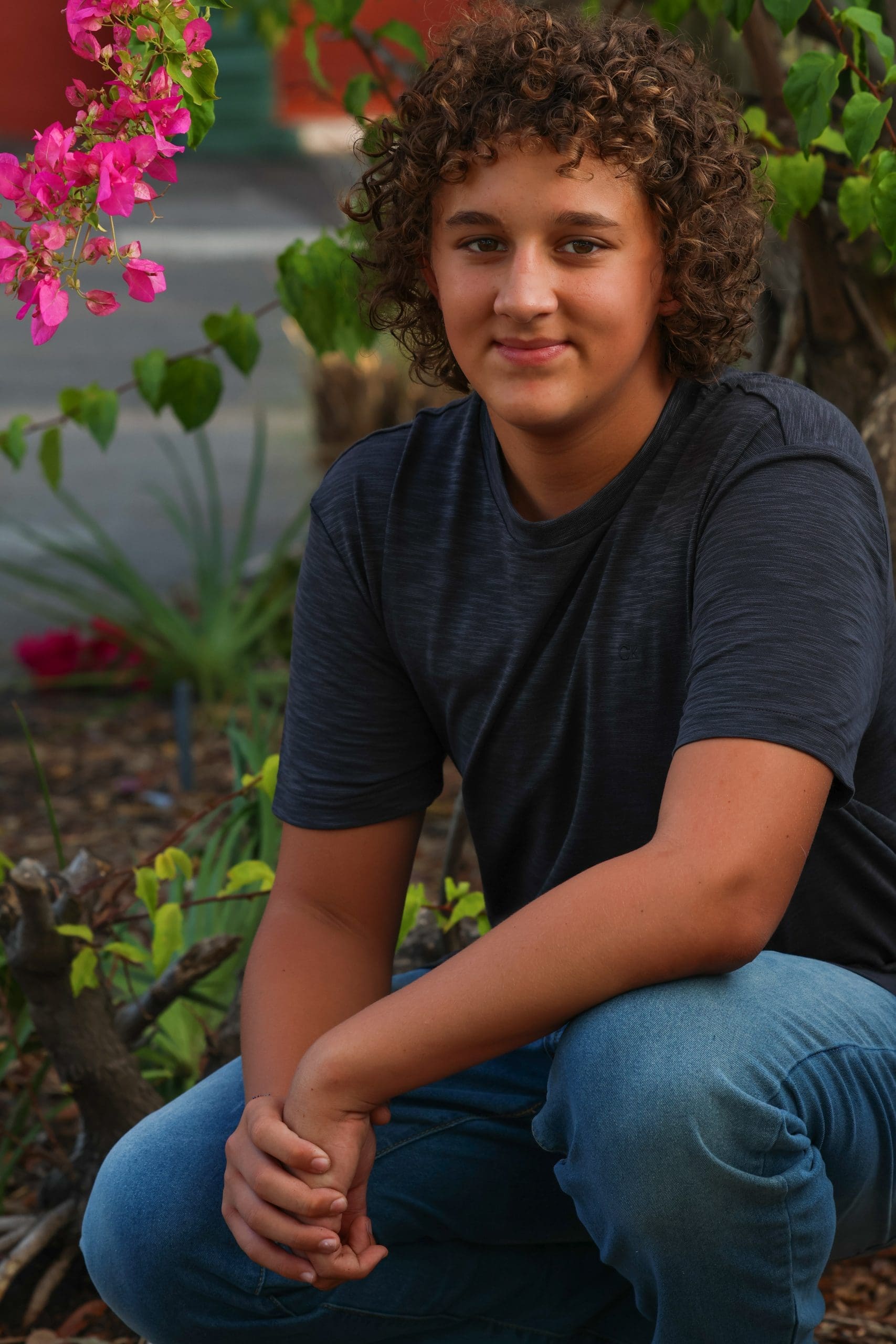 High school senior boy squatting in front of bright pink flowers in a garden, wearing jeans and a grey shirt in a casual outdoor portrait.