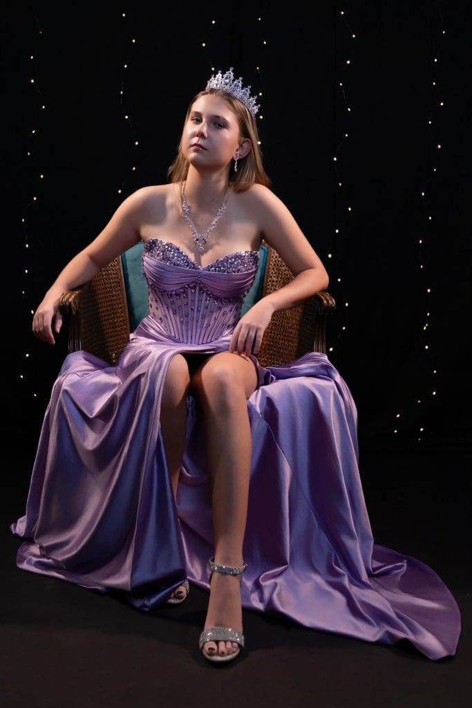 Kahlen16thBdayPortrait Studio fine art photography: Kahlen in a purple corset gown with a crown, posed in an antique chair on a black background.