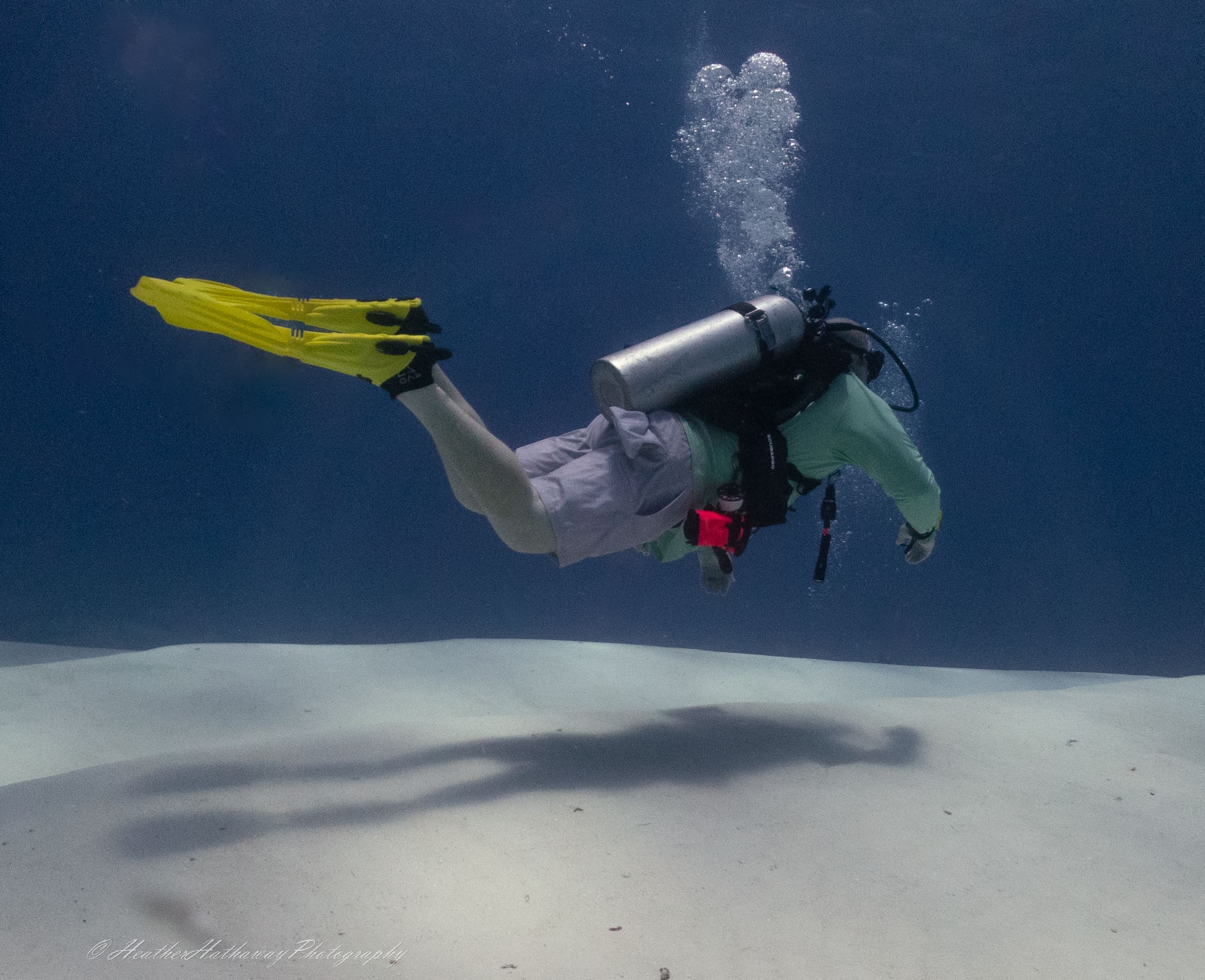 Underwater photo of a scuba diver with yellow fins swimming over white sand, bubbles trailing behind, with his shadow visible below.