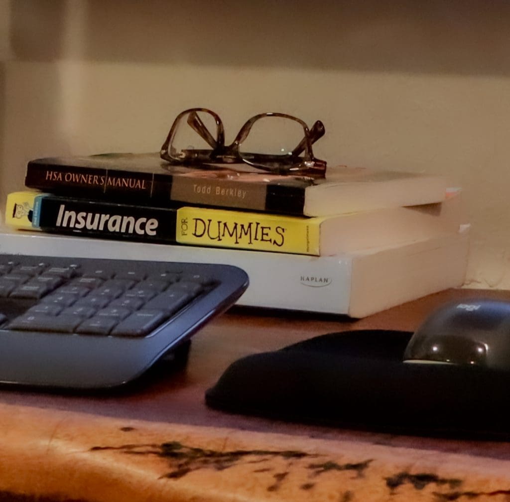 A stack of Insurance books with glasses on top sits on a desk next to a keyboard.