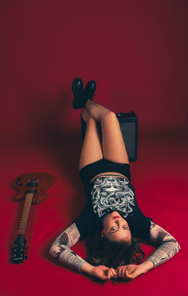 Alicia and Guitar in Red Musician Alicia poses laying on a red seamless backdrop paper with her guitar to her left.