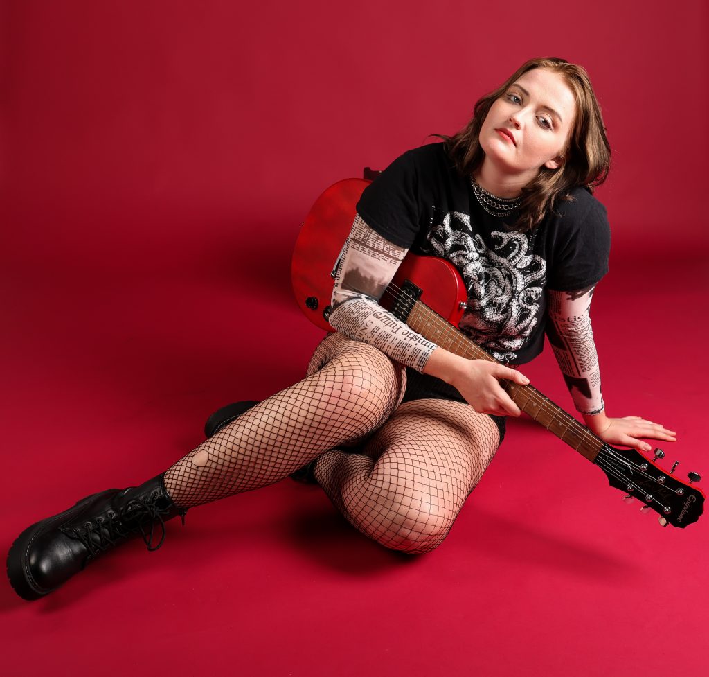 Musician Alicia Neil poses with her guitar on a red seamless paper backdrop.