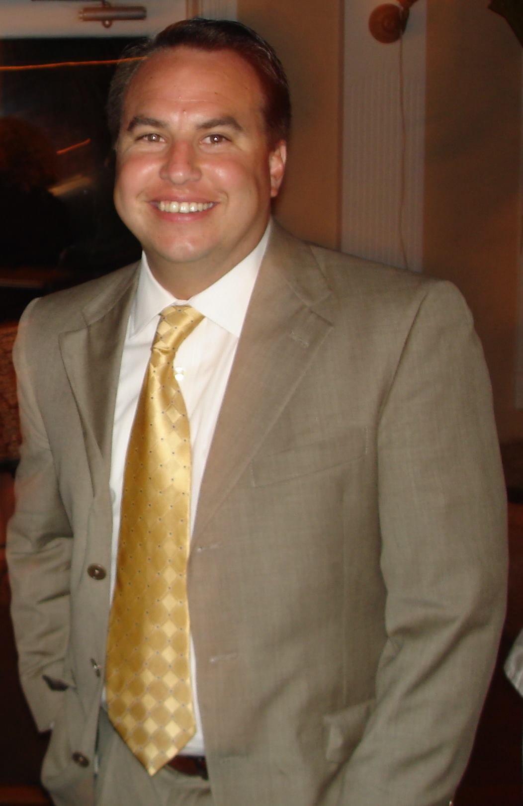 George in Suit George Noble stands waist up in a tan suit with yellow tie.